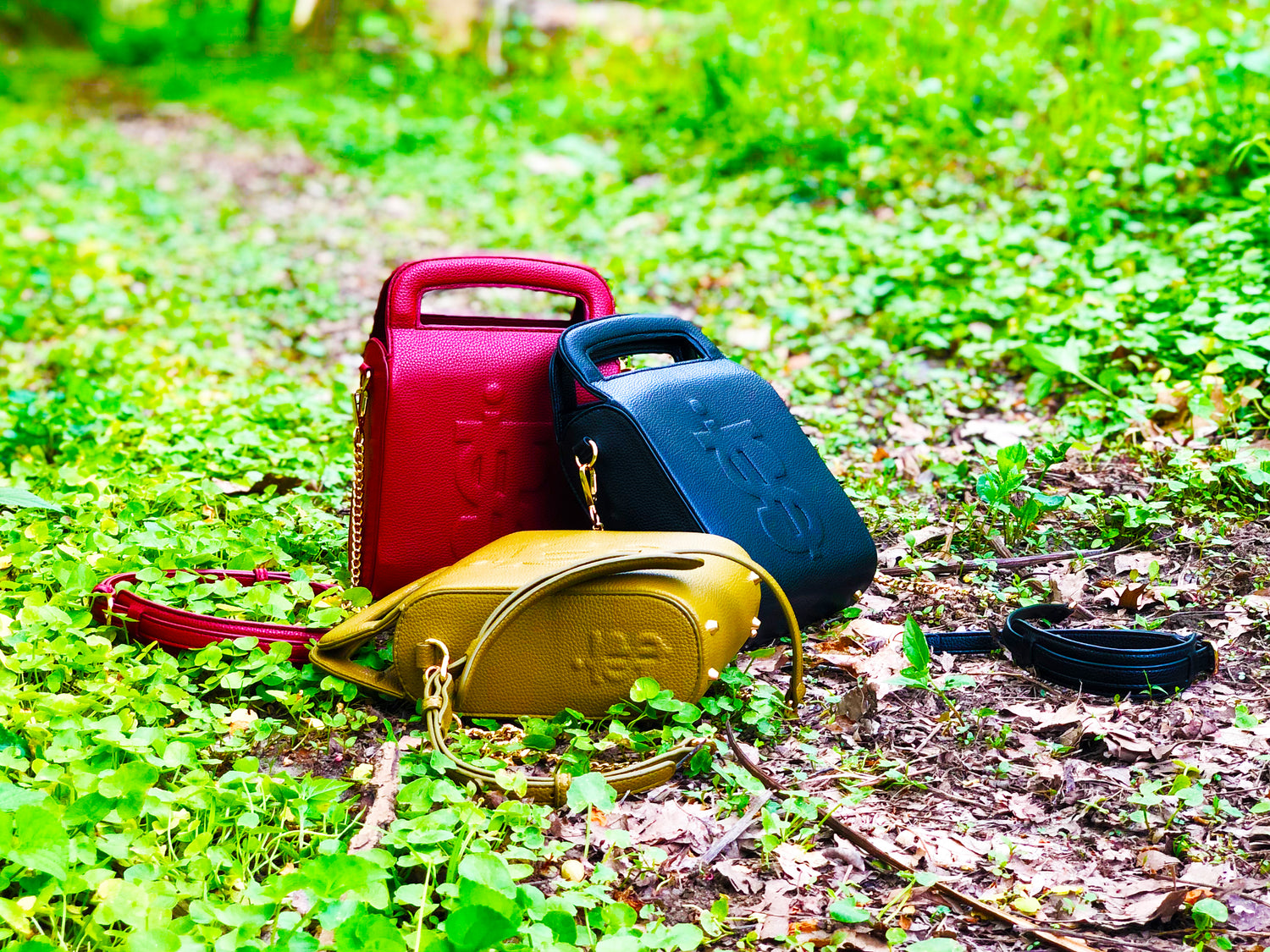 3 Black owned handbags. Oval shaped purse in burgundy, black and green with handles and straps. Purses with dollar sign I.T.S embossed on front and side stacked in a triangle on the ground of Cobbs Creek woodlands in Philadelphia, PA.