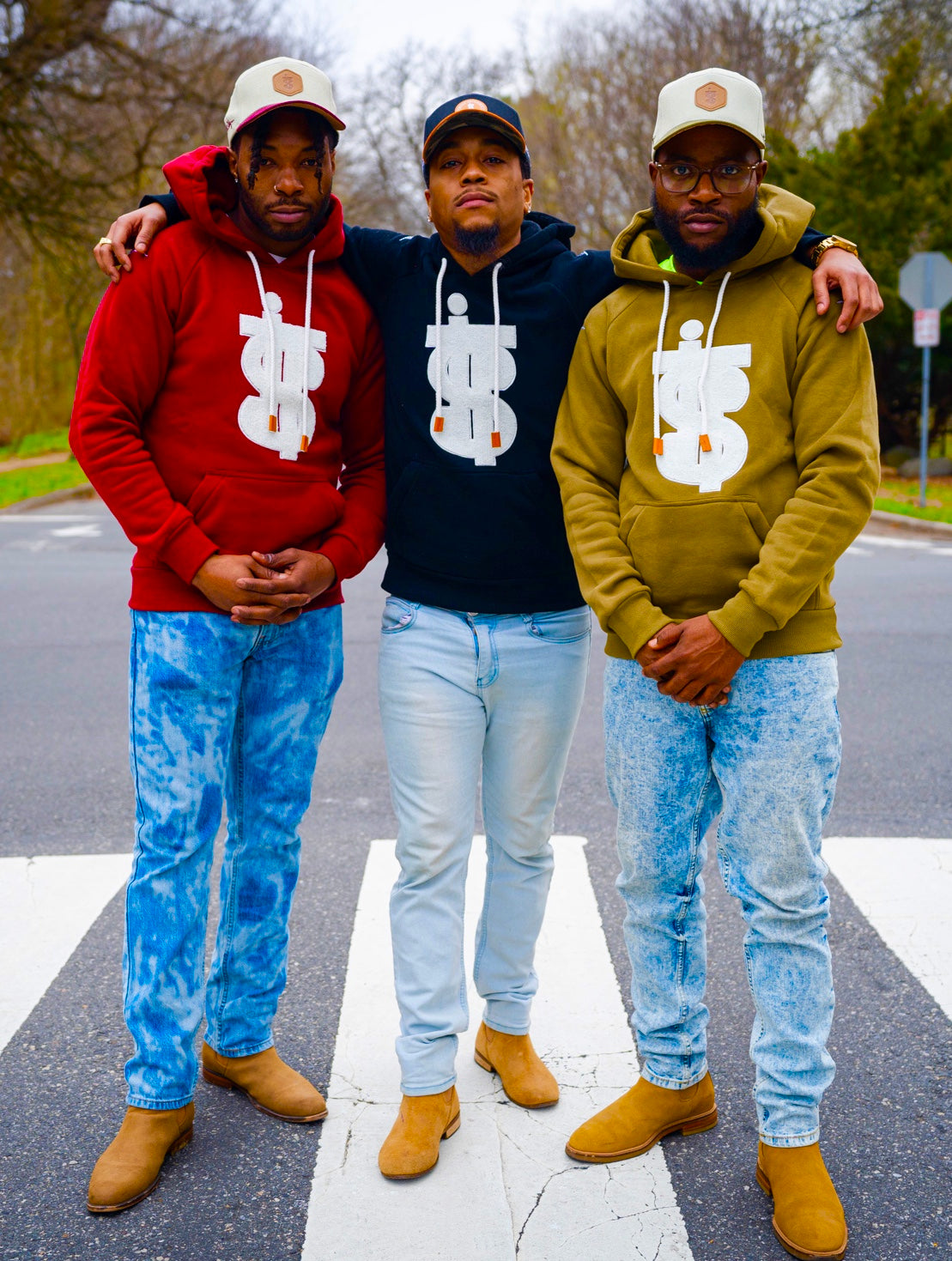 Three Black men wear I.T.S on burgundy, black, and green hoodie making a dollar sign with matching snapbacks on heads. Finishing the look with light wash jeans and Chelsea boots.